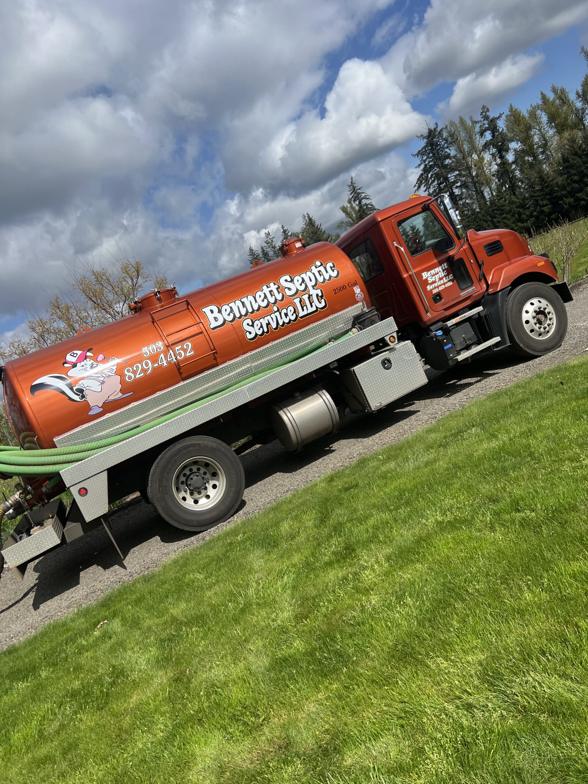 A red Bennett Septic Service LLC truck is parked on a gravel road next to green grass, with trees and clouds visible in the background.