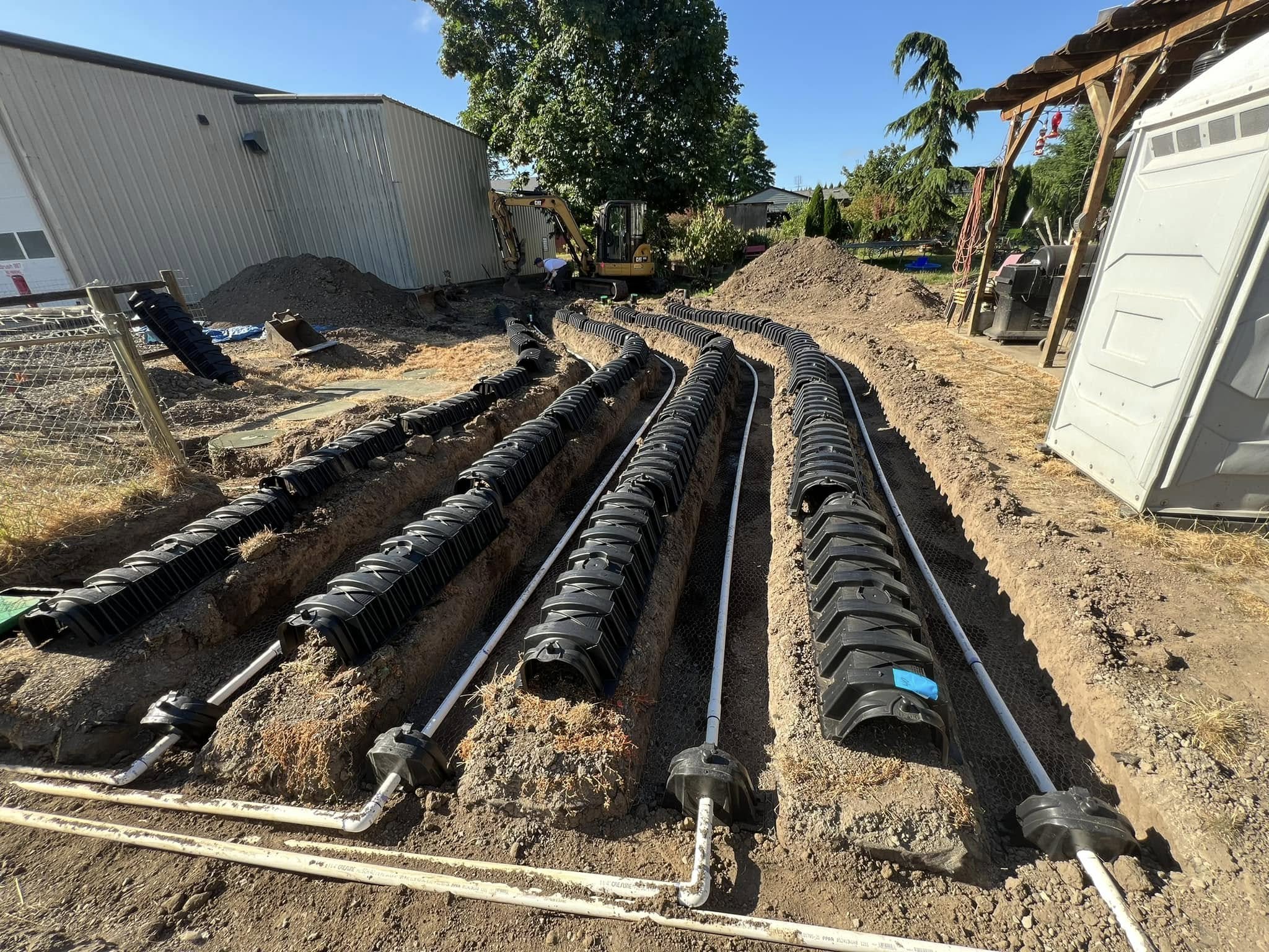 A partially installed septic drain field with multiple black plastic chambers and white pipes, surrounded by dirt and construction equipment.