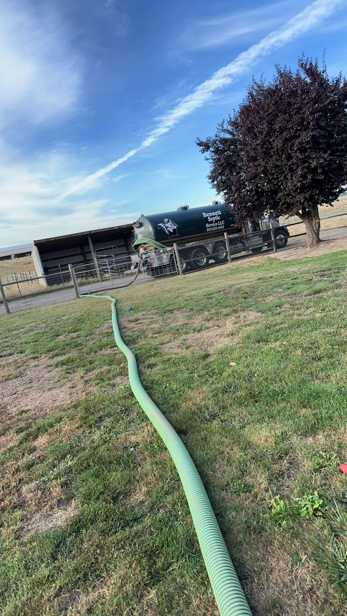 A long green hose extends across a grassy yard toward a large truck parked near a shed and tree under a partly cloudy sky.