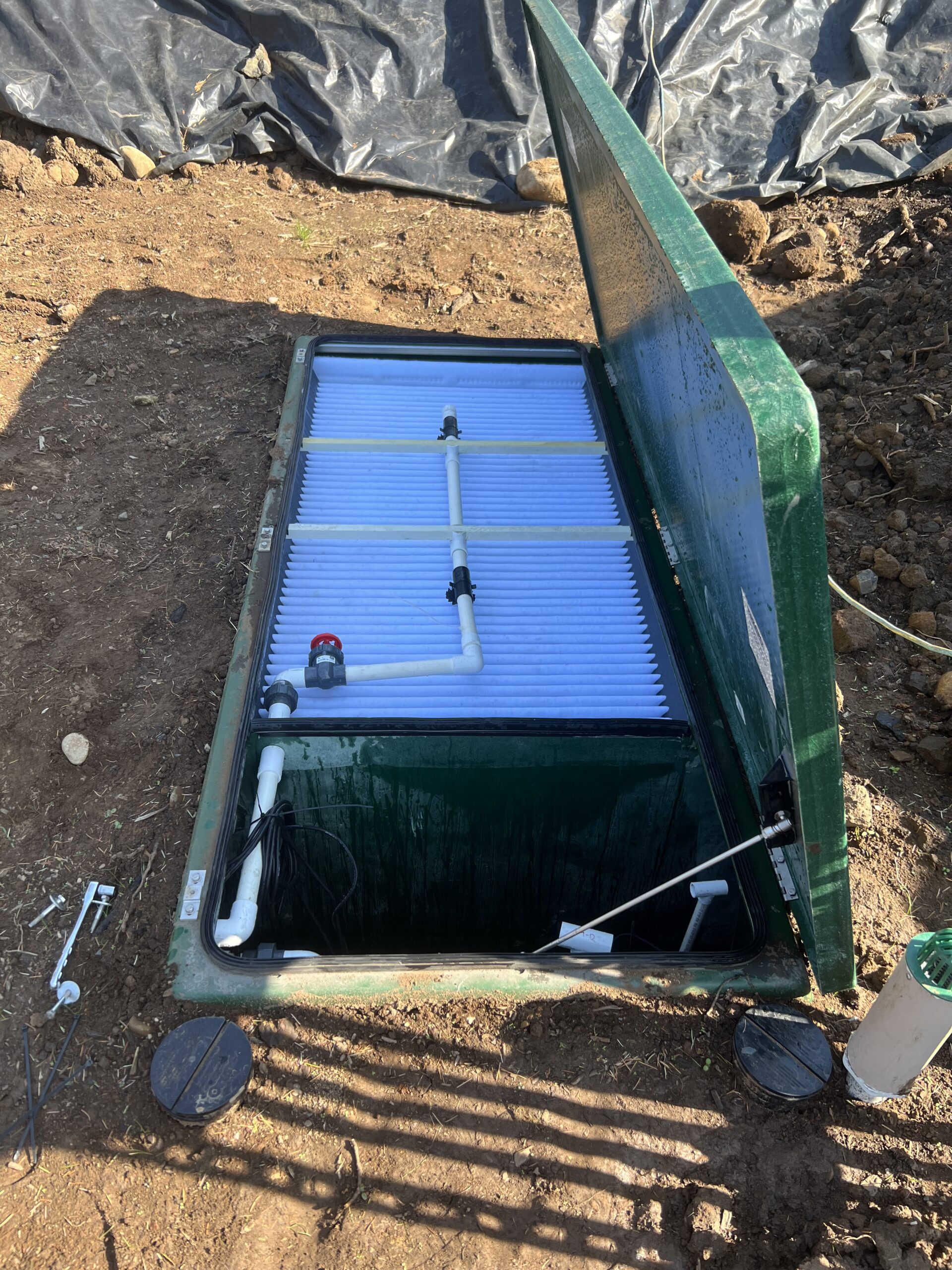A green plastic septic or water filtration system installed in the ground with its lid open, showing pipes and filter panels inside. Soil and a black tarp are visible around the unit.