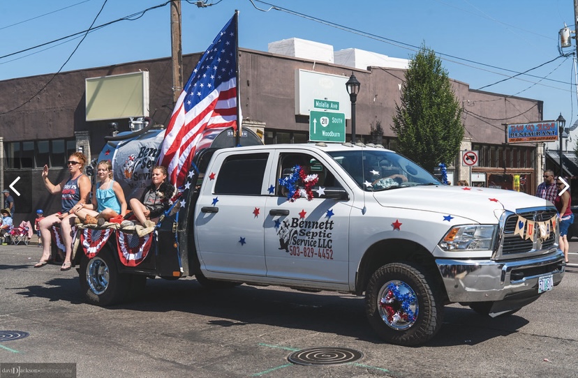 A decorated pickup truck and septic tank promoting Bennett Septic Service LLC participate in a parade, with children riding in back and American flags displayed.