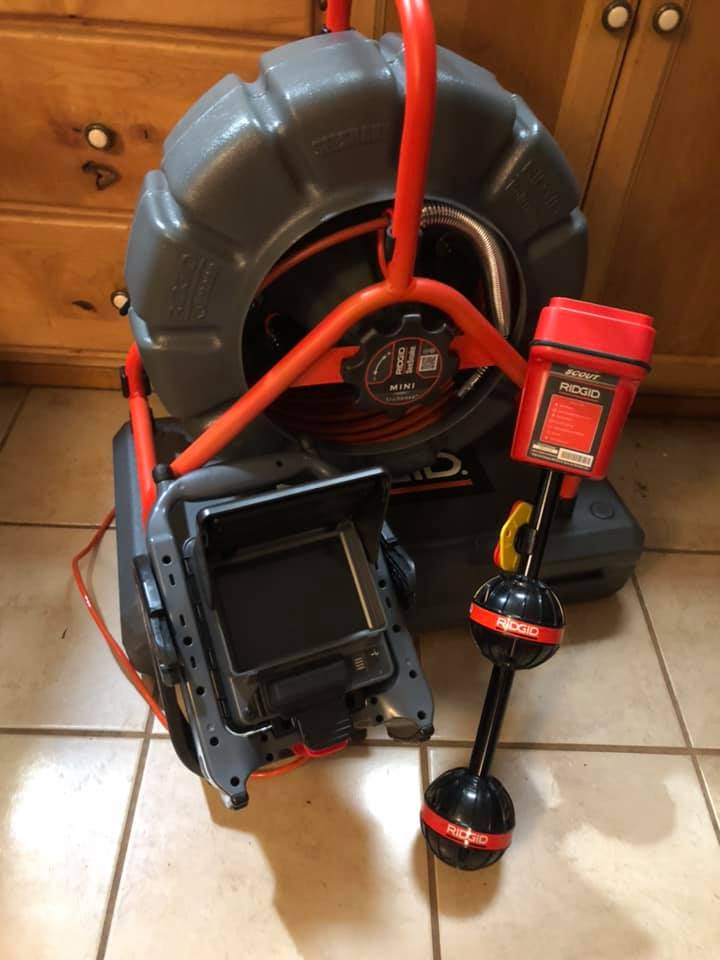A RIDGID sewer camera inspection system and locator device are placed on a tiled floor near a wooden cabinet.