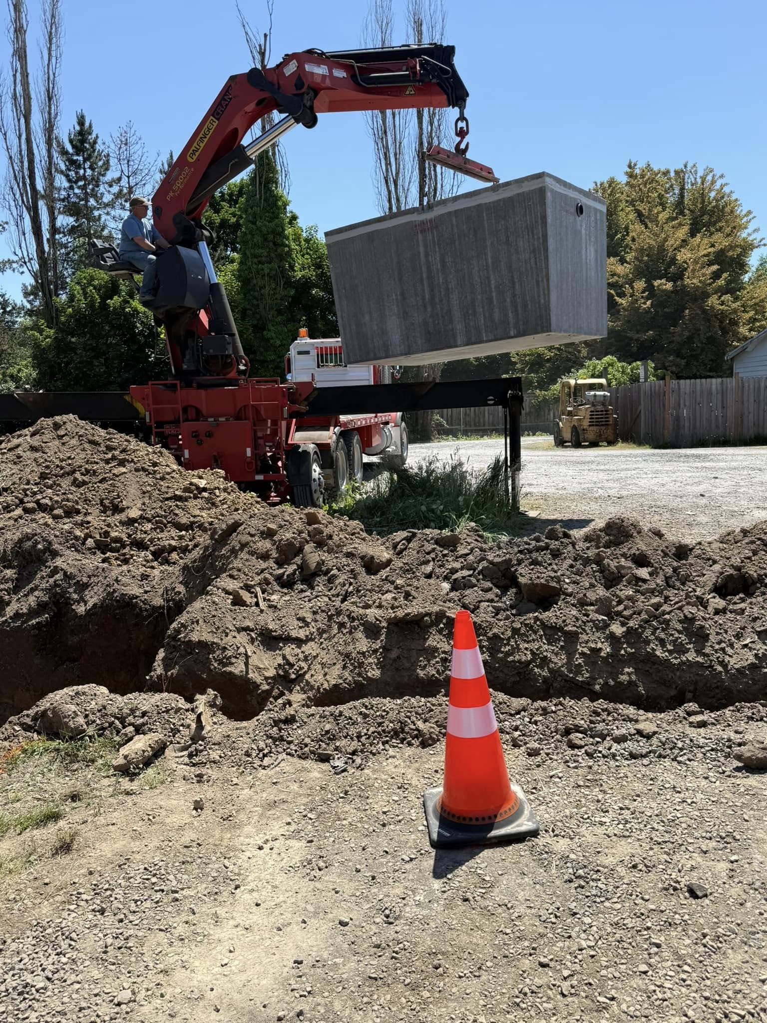 A red crane lifts a large concrete box over a dug-up area, with a traffic cone placed in the foreground on gravel.