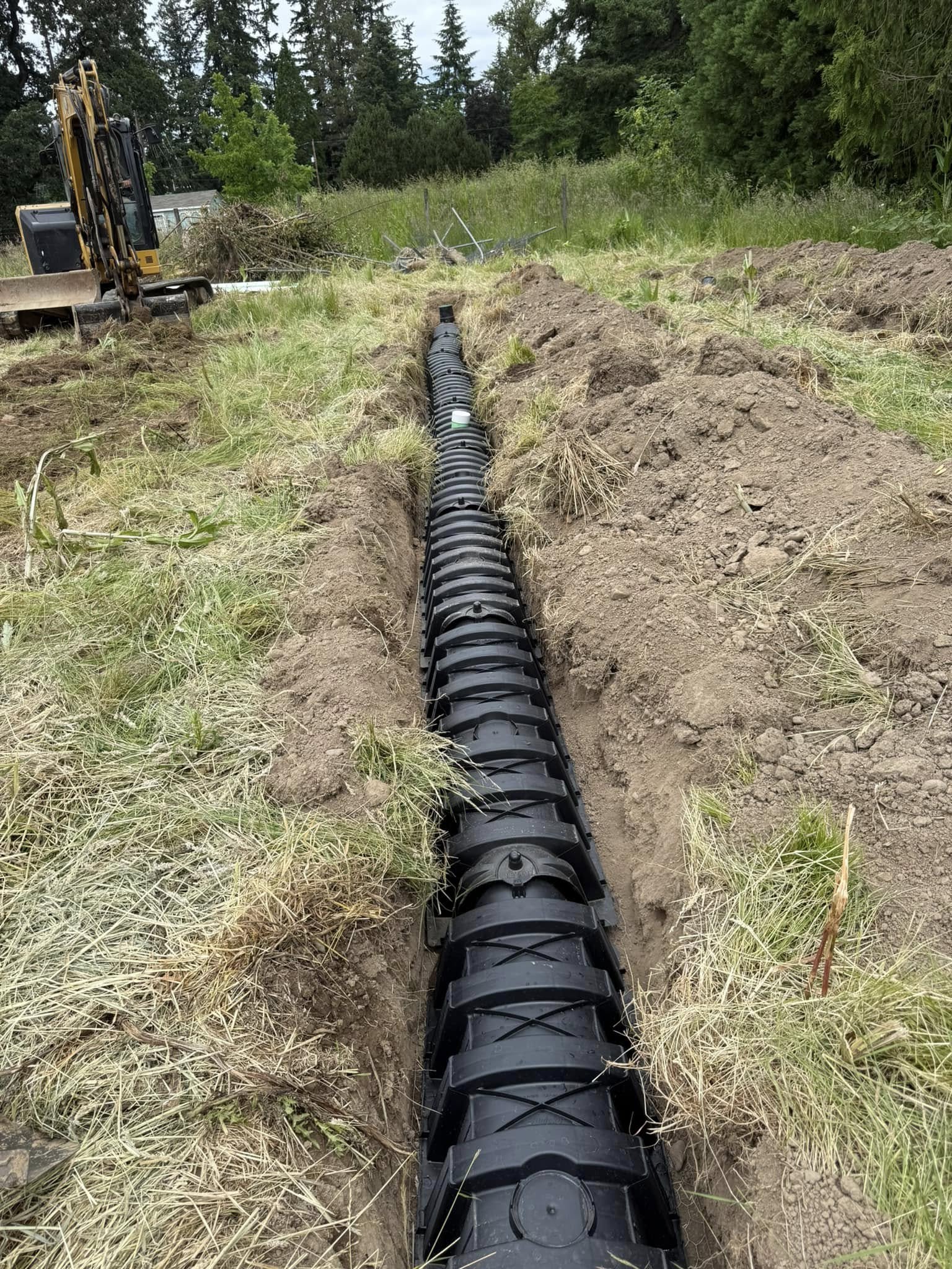 A black plastic drainage pipe is installed in a trench running through a grassy area, with construction equipment visible in the background.