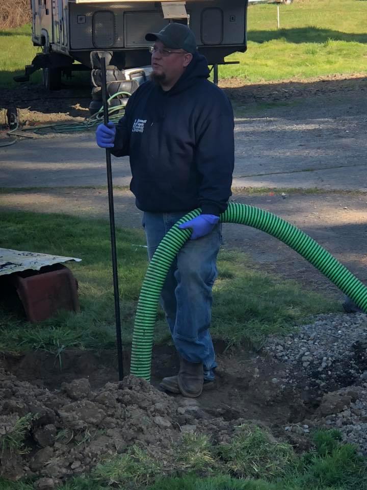 A man wearing gloves, jeans, and a hoodie stands in a dirt area holding a green hose and a metal rod, with a truck and grassy field in the background.